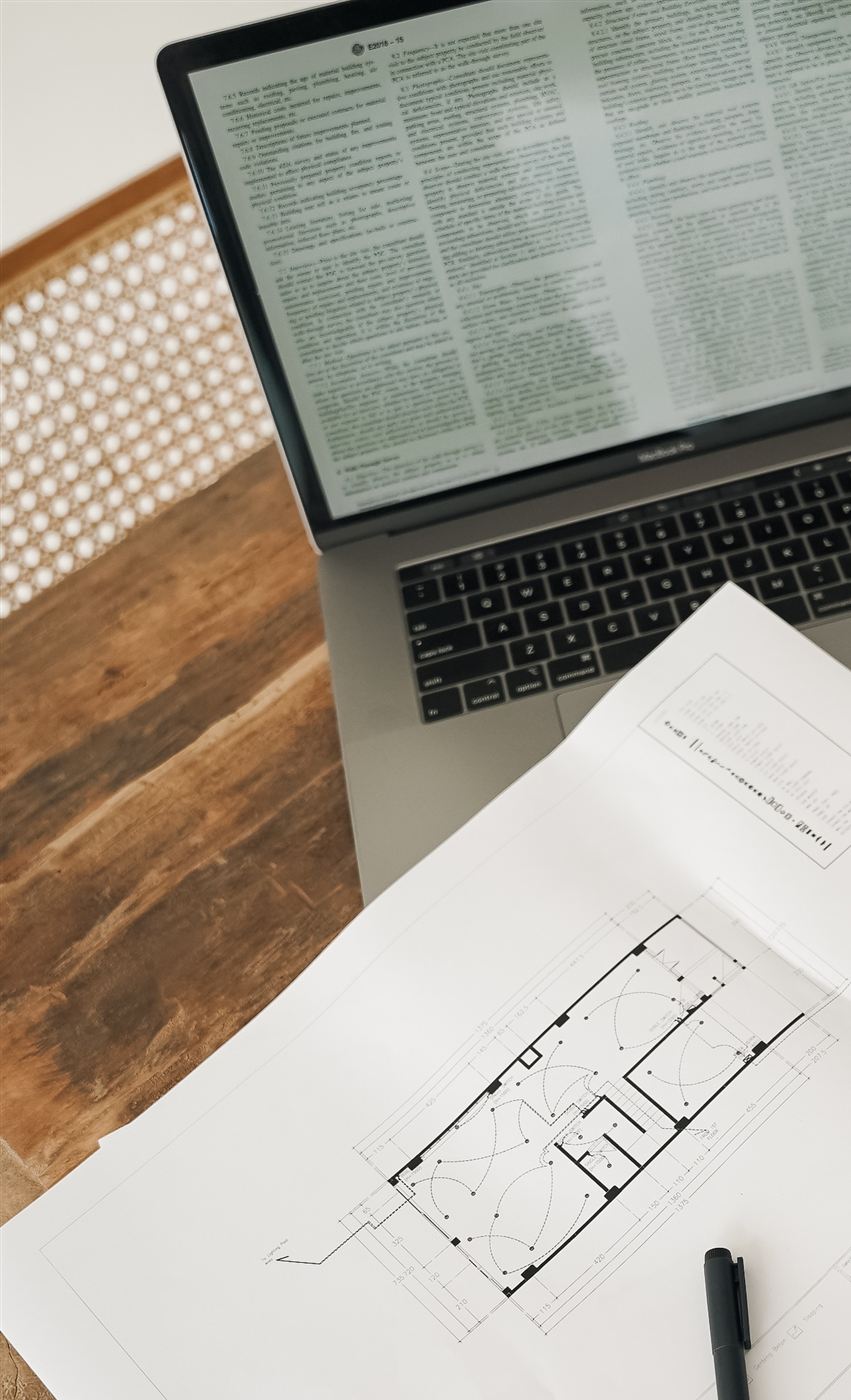 A laptop and building floor plans on a wooden desk in Bali.