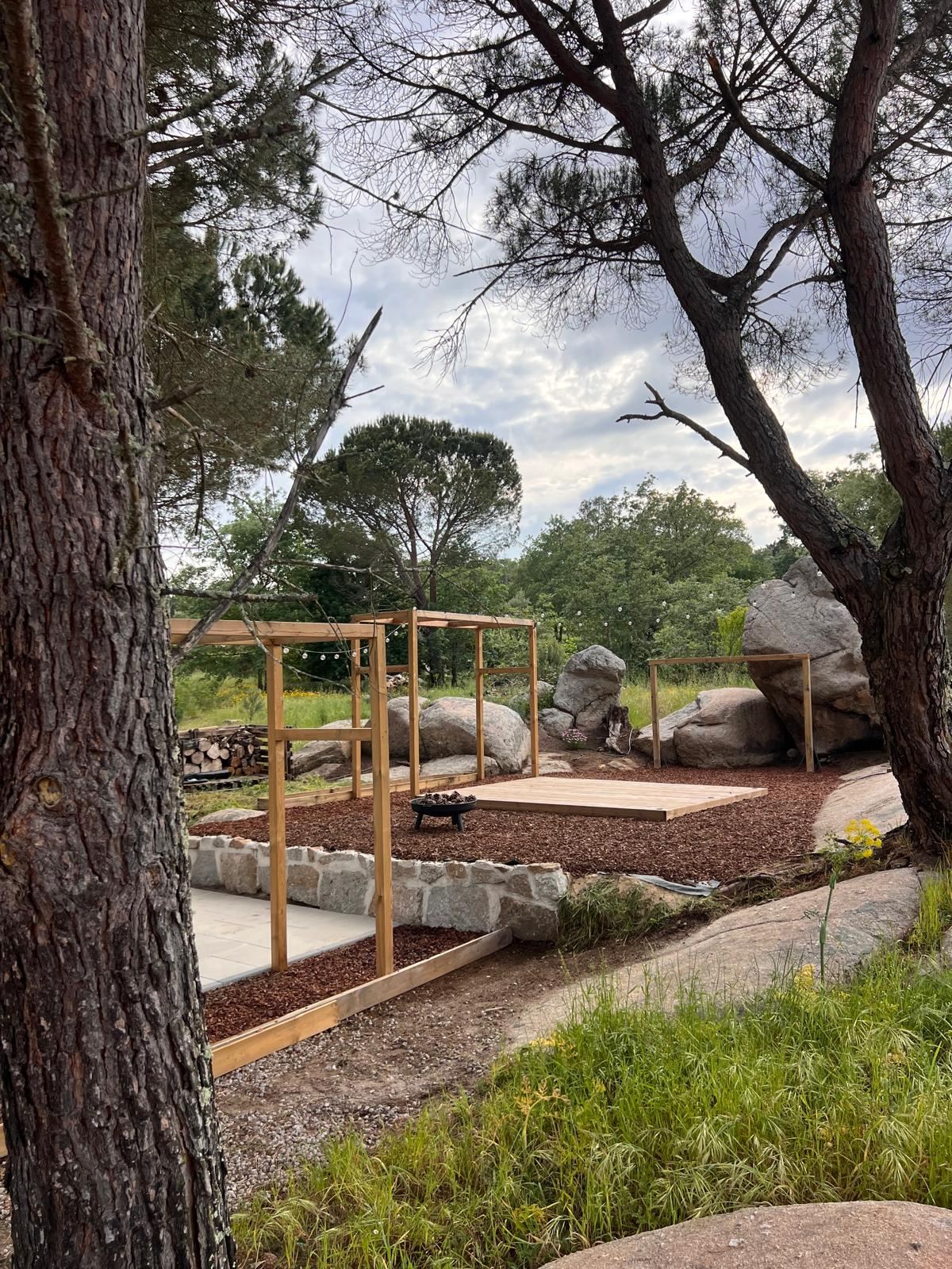 An outdoor area under pine trees, with wooden pergolas, a deck and a fire bowl between granite boulders.