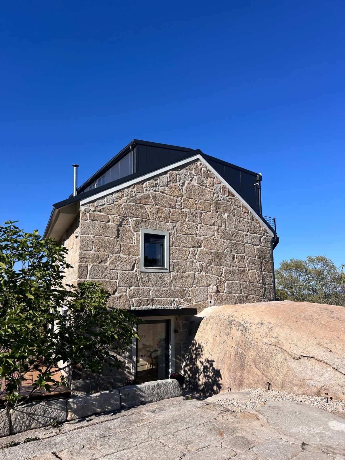 The same granite house in 2026: fully renovated, with a new dark roof, under a deep blue sky.