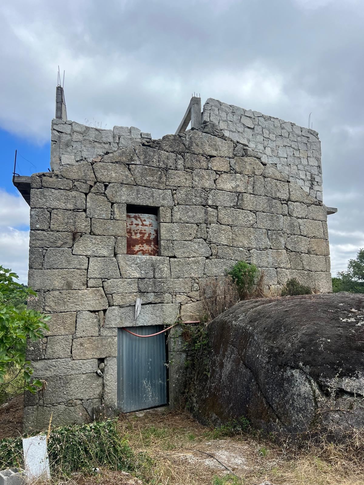 The granite main house in 2024: roofless, overgrown and in ruins, next to a large boulder.
